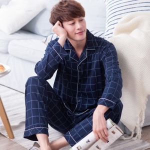 Pijama informal de hombre a cuadros de algodón azul marino de muy alta calidad llevado por un hombre sentado en una alfombra delante de una cama en una casa