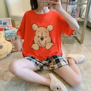Pijama de dos piezas con camiseta Winnie y pantalón corto a cuadros de moda que lleva una mujer sentada en una baldosa en una casa
