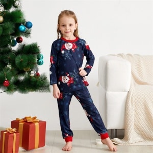 Pijama con estampado de copos de nieve y Papá Noel para la familia que lleva una niña delante de un árbol de Navidad en una casa