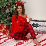 Pijama de mujer con estampado navideño y hombros descubiertos que lleva una mujer sentada en una alfombra delante de un árbol de navidad en una casa