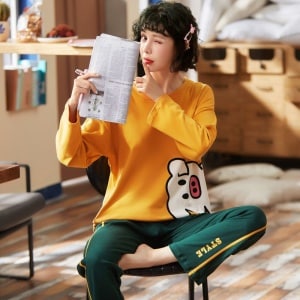 Pijama de algodón con jersey cerdito amarillo y pantalón verde que lleva una mujer sentada en una silla leyendo el periódico en una casa