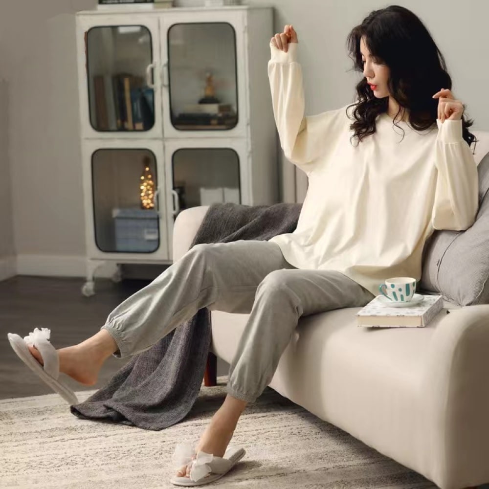 Pijama de verano con jersey blanco de mangas murciélago y pantalón gris que lleva una mujer sentada en el sofá de una casa